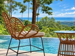 Breezy outdoor seating by pool with ocean view and lush green forest backdrop.