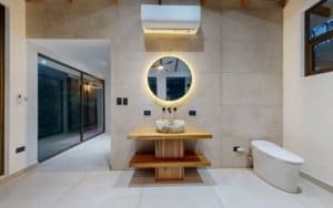 Modern bathroom with round backlit mirror, stone walls, wooden vanity and frosted glass door.