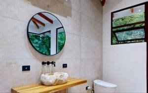 Modern bathroom with round mirror over stone sink and wooden counter, natural light.