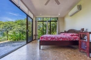 Bedroom with red bedding and glass wall overlooking lush terrace and trees.