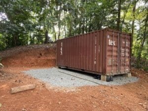 Rustic shipping container on mountain land with forest background and gravel base.