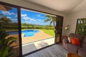 Panoramic backyard with oval pool and lush tropical landscaping seen from interior living room.