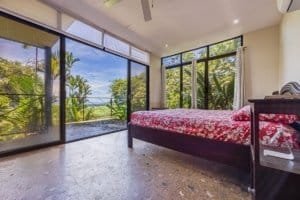 Panoramic bedroom with large glass doors to tropical balcony and ocean view.