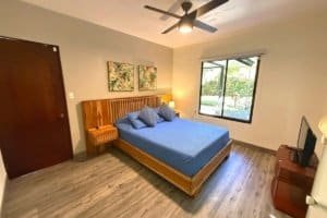 Bedroom with king bed, blue linens, wooden furniture, and a view of a garden through the window.