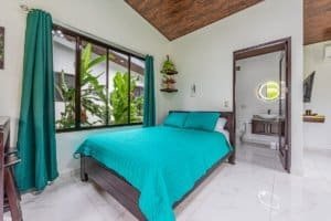Bright bedroom with teal bedding and large window overlooking tropical plants.