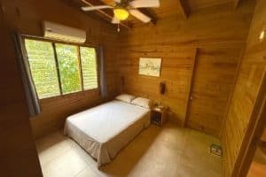Interior of a rustic jungle hotel room with wooden walls, a bed, and a window showing greenery.