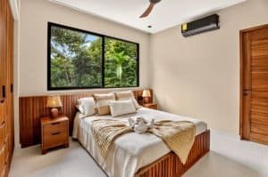 Master bedroom with large window overlooking lush tropical jungle and wooden furniture.