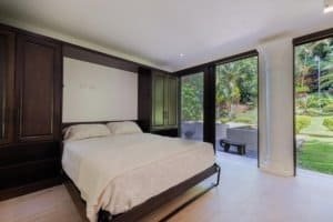 Spacious bedroom with large windows overlooking lush garden and outdoor patio.