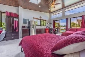 Bright bedroom with burgundy bedspread and modern kitchenette in open-plan villa.