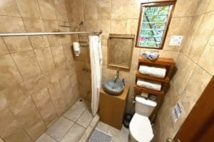 Small bathroom with stone sink, toilet, window, and wooden shelves in Costa Rica.