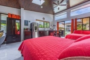Bright bedroom with red quilted bedding, modern kitchenette, and large windows in a tropical villa.