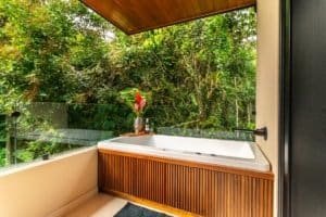 Outdoor balcony bathtub with glass railing and lush tropical greenery surrounding.