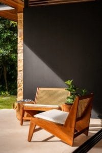 Outdoor wooden lounge chairs with cushions on a shaded patio by a dark wall and greenery.