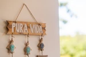 Pura Vida wooden sign with hanging shells and beads on a wall.