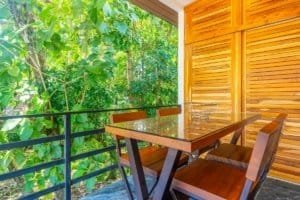 Oceanview balcony with wooden table and chairs surrounded by lush trees.