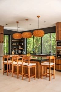 Modern kitchen with island and wooden bar stools open to lush jungle view.