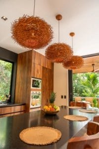 Luxurious tropical kitchen with woven pendant lights and wood cabinetry, overlooking lush greenery.