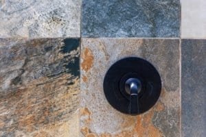Close-up of a round black shower valve on multicolored slate tiles.
