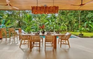 Tropical outdoor dining table set under a shaded patio with lush jungle garden.