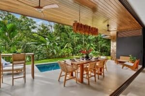Outdoor dining area with wooden table and chairs near pool, surrounded by lush tropical plants.