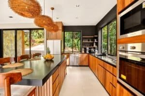 Modern jungle kitchen with wooden cabinets, black countertops, and large windows.