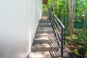 Concrete outdoor stairs with metal railing beside a white wall and greenery.