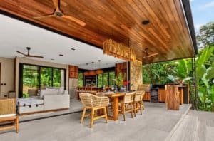 Outdoor kitchen and dining area with wooden ceiling, gray tiled floor, and lush tropical greenery in.