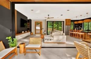 Open-plan living room with sofa, wicker chairs, modern kitchen, and tropical greenery view.