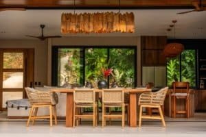 Luxurious tropical dining area with wooden table, woven chairs, and natural light from large windows.