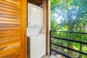 Compact stacked washer dryer on balcony with wooden walls and lush green plants outside.