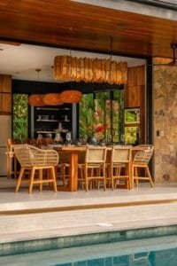 Open-air dining area with wooden chairs, stone wall, and swimming pool in tropical jungle setting.