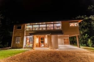 Night view of a modern two-story jungle home with stone entry, illuminated windows, and surrounding.